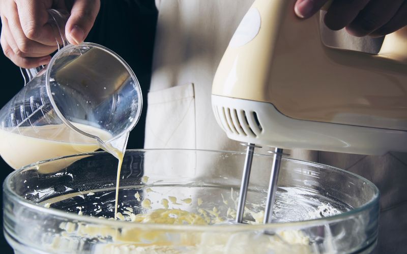 A mixing bowl with a person adding milk to it and mixing it with a hand mixer